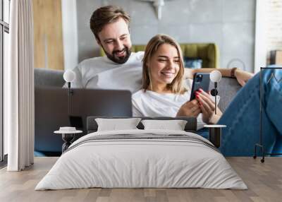 Beautiful young couple relaxing on a couch at home using laptop computer and mobile phone Wall mural